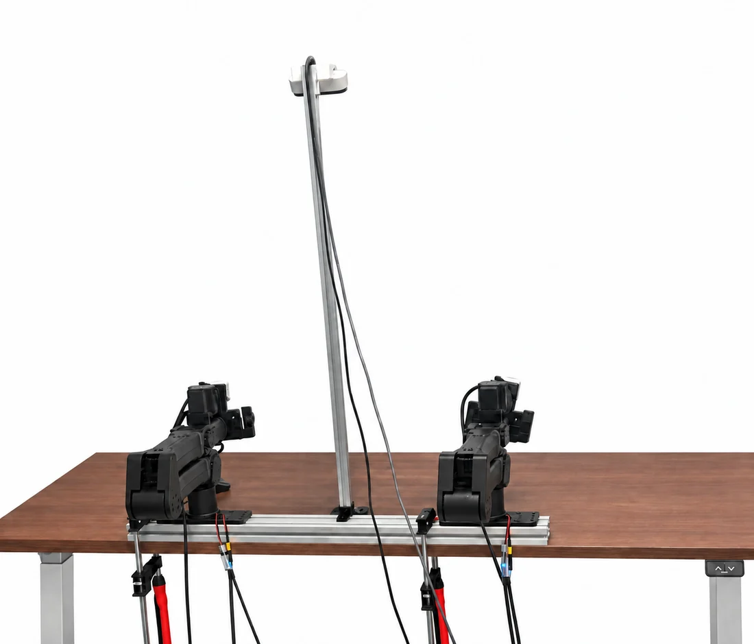 Photograph of two robot arms bolted to an aluminium rail that lies on a wooden tabletop, facing each other, with an overhead camera on a mast between them and cabling running down past the table edge.
