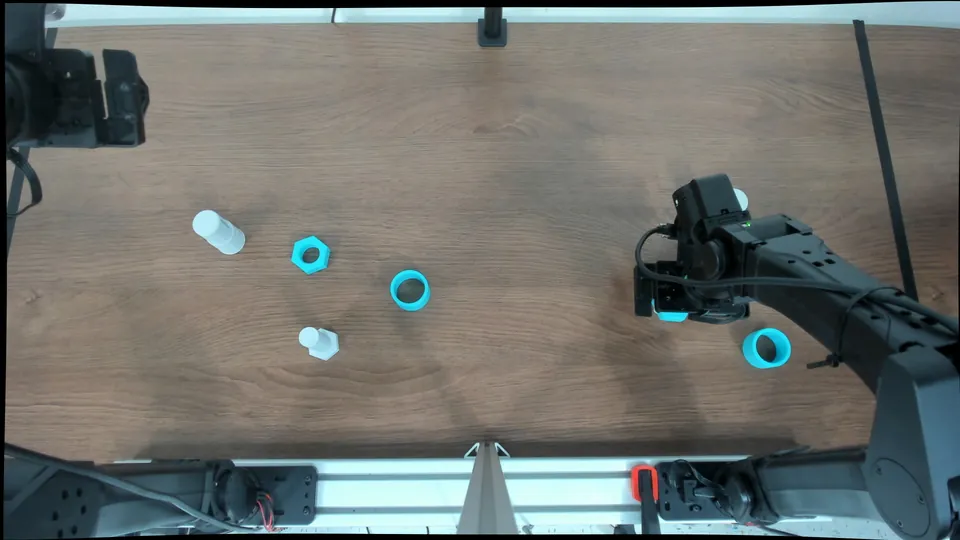 Overhead camera frame of a wooden tabletop with four 3D-printed parts lying apart on it, a white cylinder, a teal hexagonal nut, a teal ring and a white hexagonal nut, one robot arm reaching over the table and the base of a second arm visible.