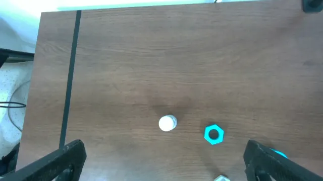 Wrist-camera view looking down at the wooden tabletop, the two dark gripper jaws at the bottom edge of the frame, a white cylindrical post and a teal hexagonal nut lying on the table ahead of them.